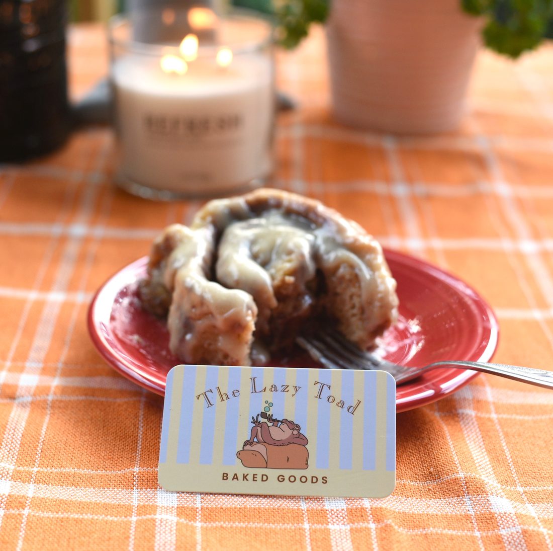 A frosted cinnamon roll on a red plate beside a Lazy Toad business card