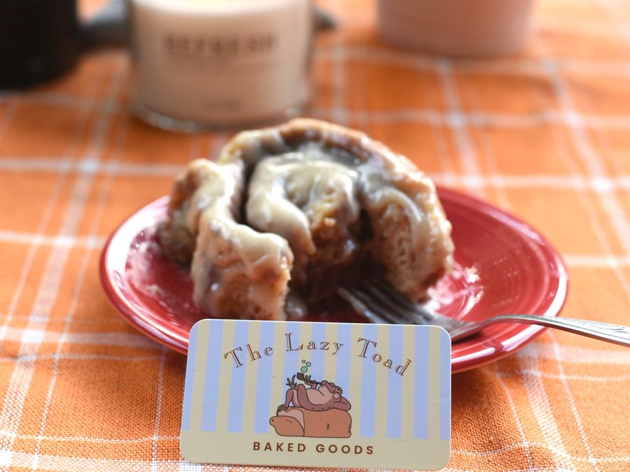 A frosted cinnamon roll on a red plate beside a Lazy Toad business card
